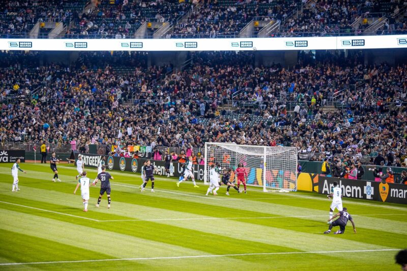 The Oakland Roots playing off against a team with a black kit on the soccer/football pitch. Players watch as the ball falls from the sky, into the penalty box.