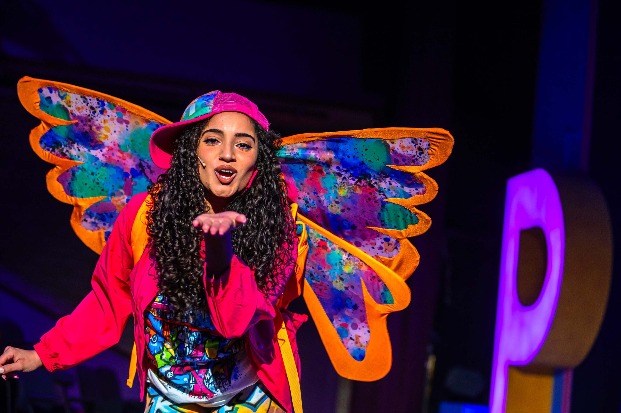 A woman on stage, wearing butterfly wings, blows a kiss to the audience.