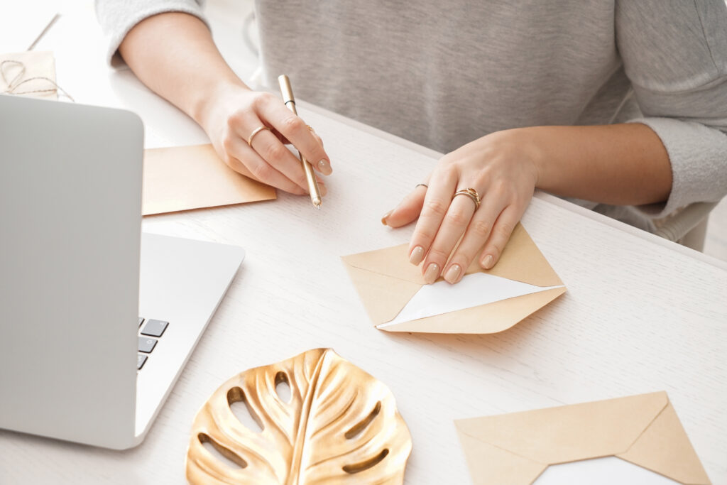 woman sitting in front of laptop writing on beige thank you cards with a pen in her right hand and a gold leaf decoration on her desk