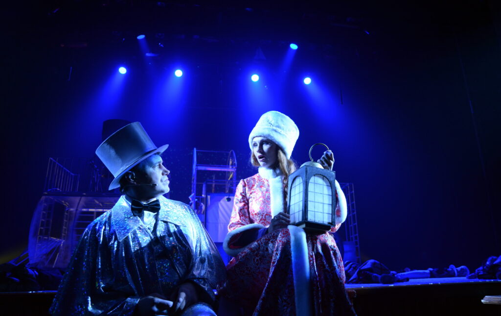 An actor and actress dressed in old-timey winter clothes and holding a lantern in a Christmas musical