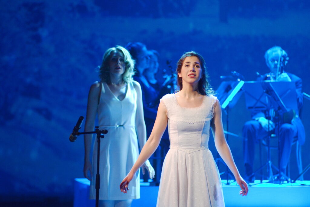 two women in white dresses against a blue-lit background, singing as part of a performance